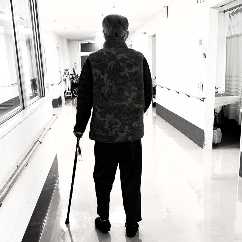 回復期病棟の廊下で杖を使って歩行訓練を行う後ろ姿